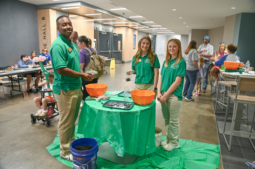 Three young people wearing green polos and standing around a table.