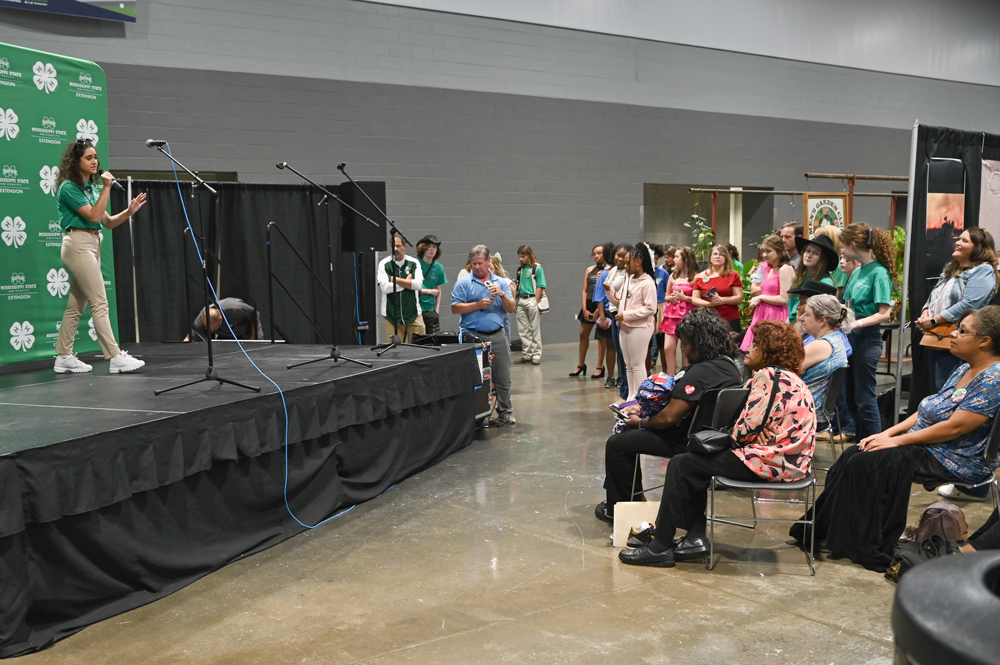 : A young woman holding a microphone is standing on a stage. A 4-H banner is in the background.