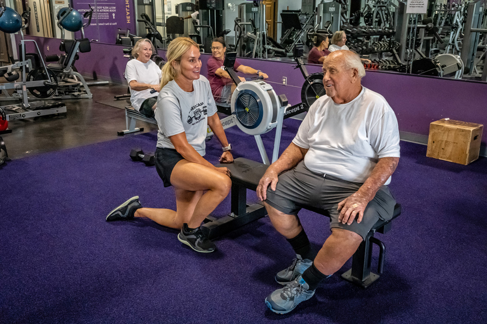 Two people in a gym; one is sitting on a weight bench, while the other is kneeling on one knee beside the bench with a hand resting on it for support.