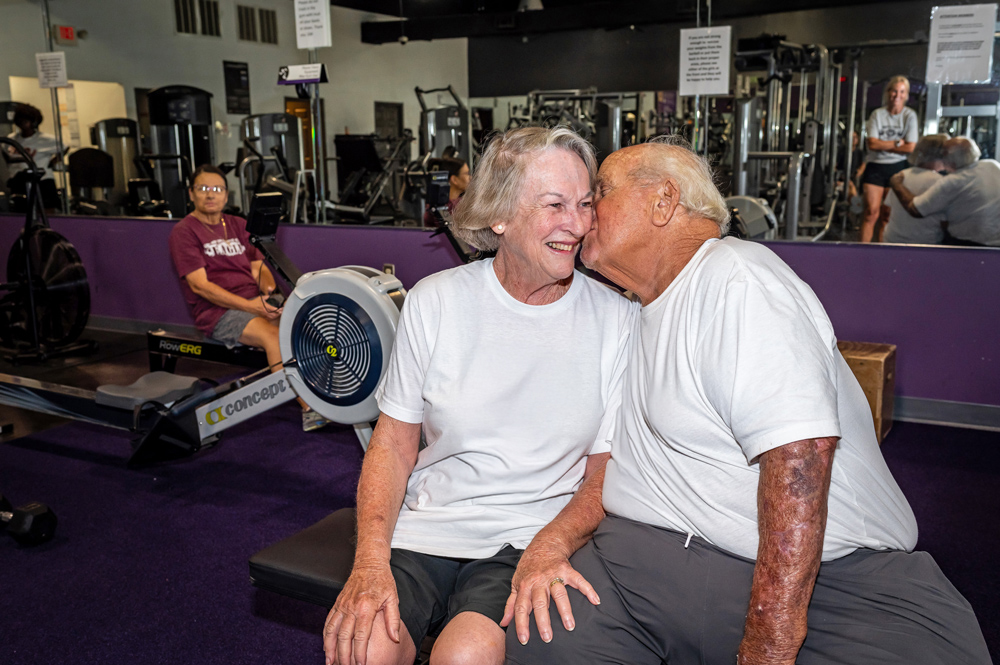 A couple sitting together in a gym; one person gently rests their face against their partner’s face.