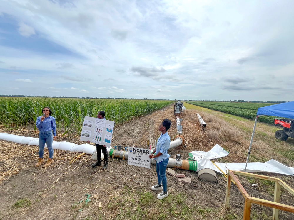 A person with a microphone speaks in a farm field, corn on the left and soybeans on the right, while another person holds a poster and a third person looks on.