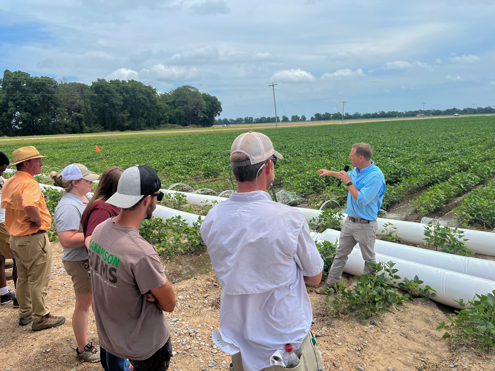 A person speaking with a microphone stands in front of a polypipe with water being released into rows of soybeans, while a group of people watches.