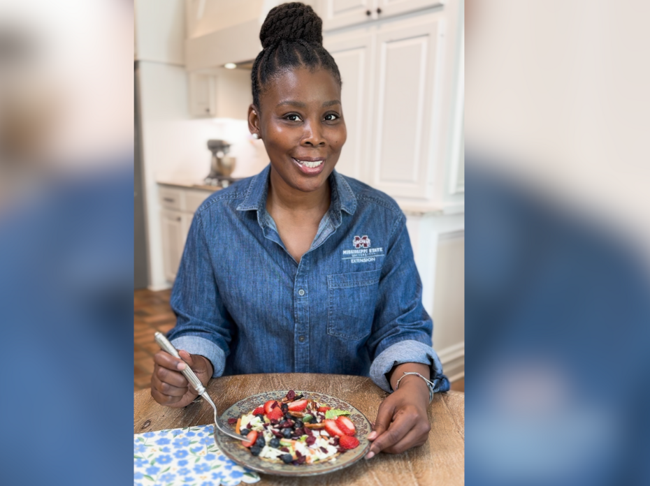 A woman sits at a kitchen table with a bowl of yogurt and berries.