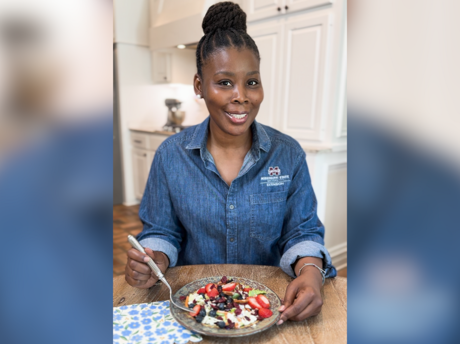 A woman sits at a kitchen table with a bowl of yogurt and berries.