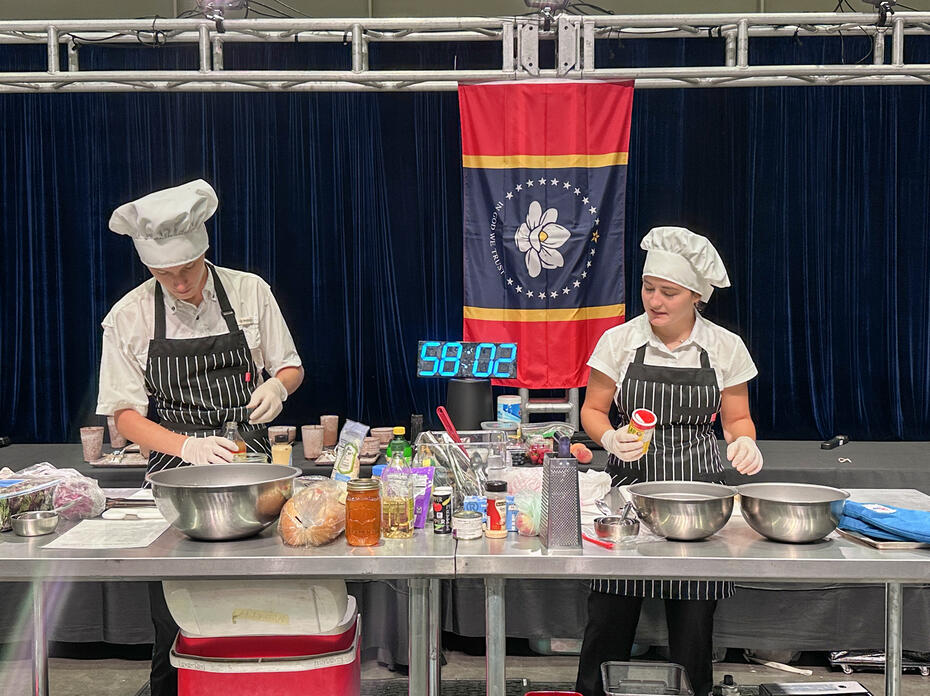 Two people prepare food during a cooking competition with a large timer in the background.