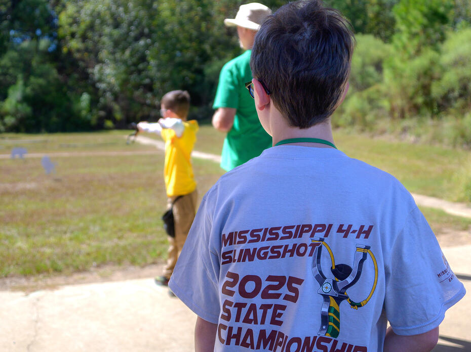 In the foreground, a boy pictured from behind wearing a T-shirt reading “Mississippi 4-H Slingshot 2025 State Championship” looks on as another boy aims at a target with a slingshot.