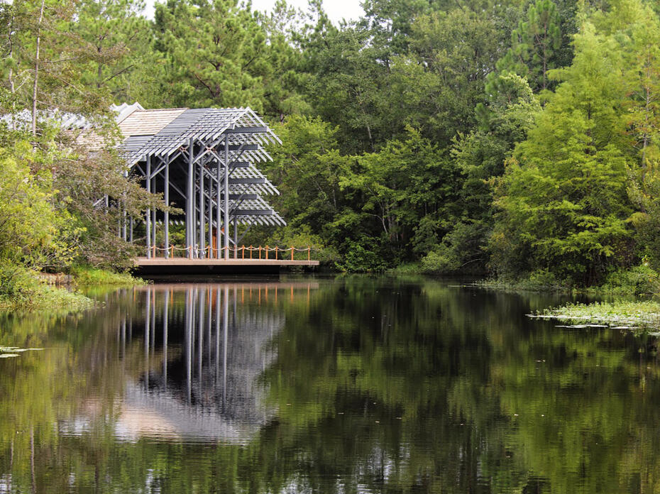 The Pinecote Pavilion, a symmetrical, wooden shed with exposed beams and a broad gable roof, stands across the pond at the Mississippi State University Crosby Arboretum. 