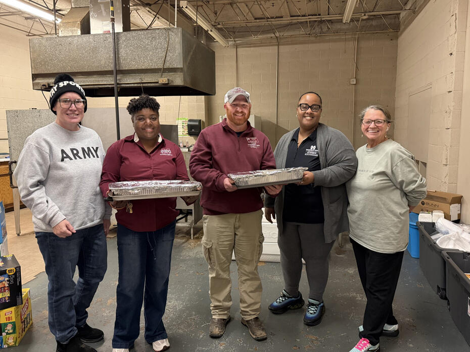 A group of five people standing together in an industrial-style room, with two of them holding large foil-covered food pans. Boxes, supplies, and equipment are visible around the room.