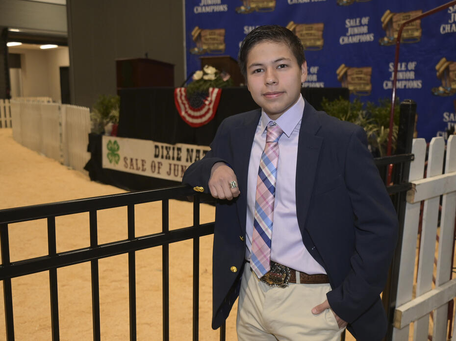 A person in a blazer and tie leans on a fence inside the Dixie National Sale of Junior Champions arena.