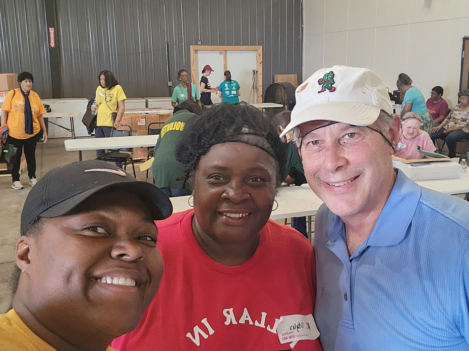 Three people pose for the camera in a church auditorium with others in the background as they prepare to distribute food.