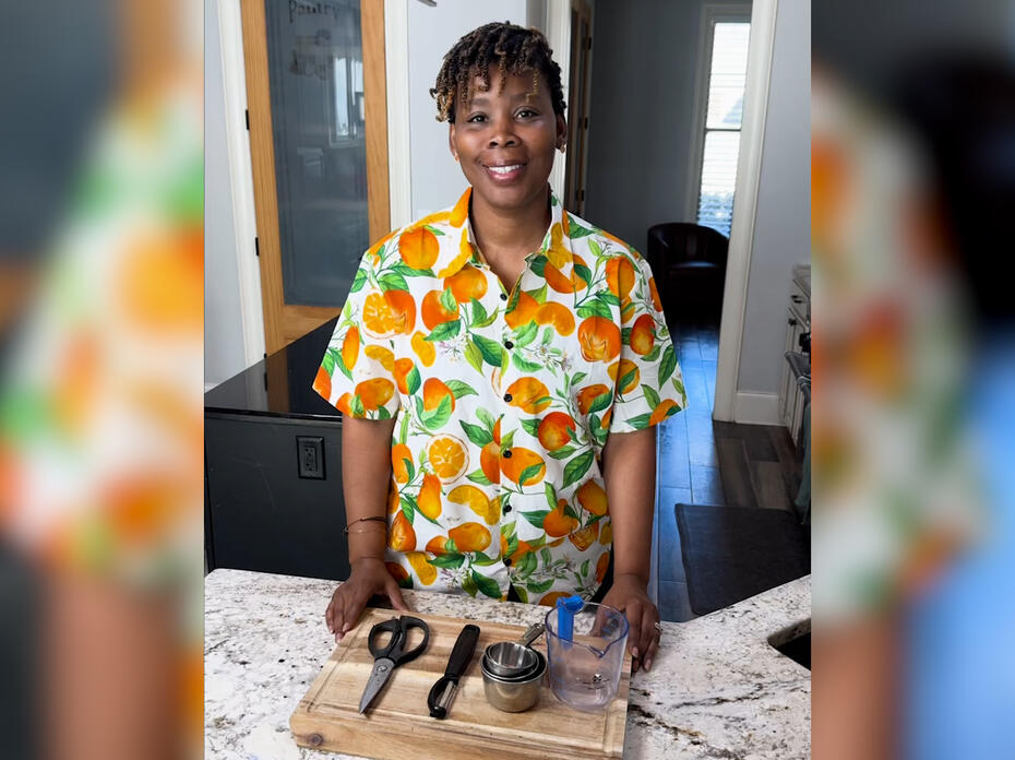 A woman stands in a kitchen with a cutting board, kitchen scissors, a vegetable peeler, and measuring cups on the counter in front of her.