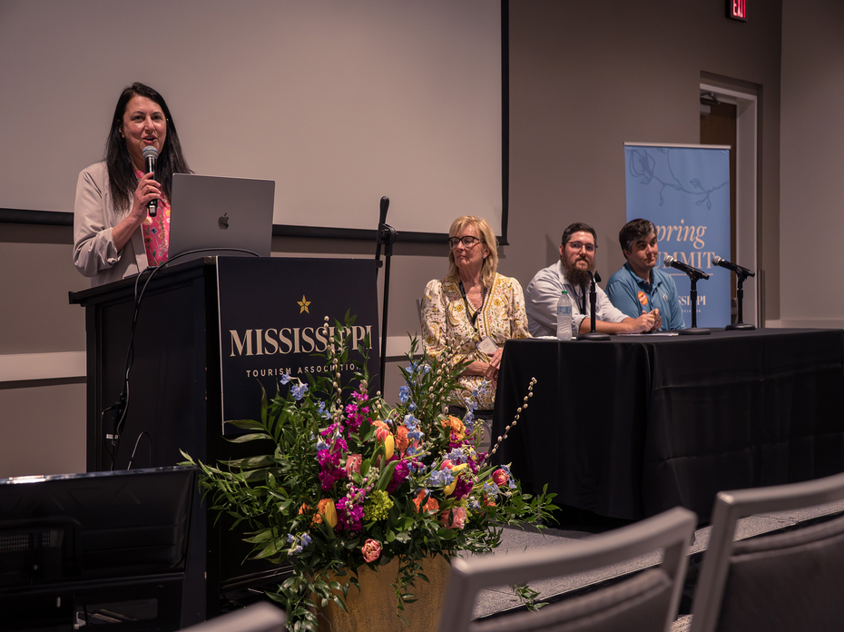 A speaker at a Mississippi Tourism Association podium addresses a panel seated behind a table in an auditorium.