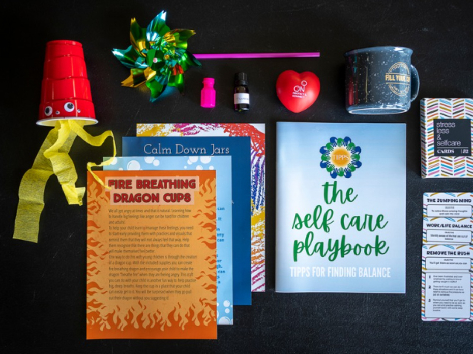 The contents of one toolkit, including a homemade fire-breathing dragon cup, various literature on parental self-care, strategies for emotion regulation and directions for crafts that support hand-eye coordination and fine motor skills, along with a coffee mug, essential oils, handheld pinwheel, a stress ball and bubbles. 