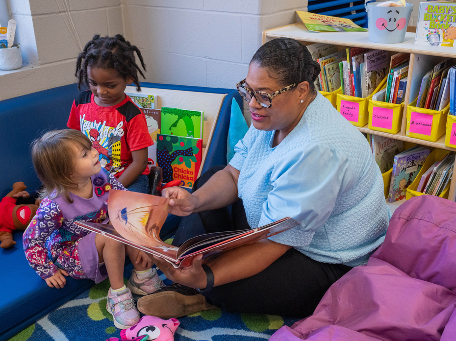 An adult reads from a picture book to two young children seated in a classroom reading corner.