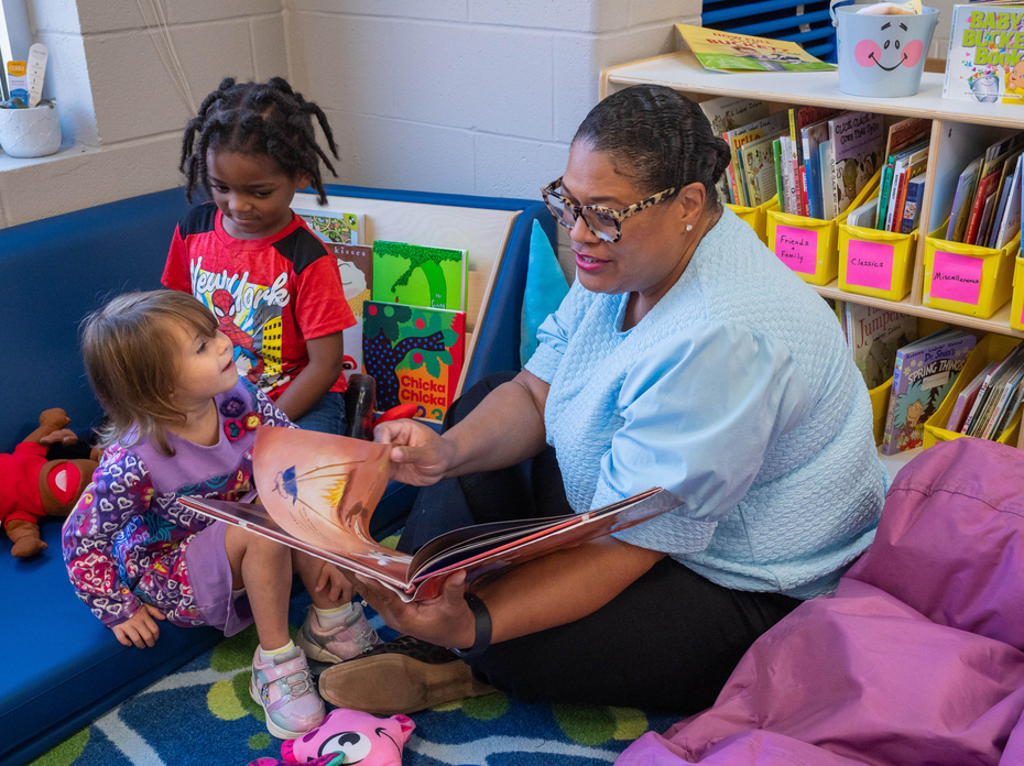 An adult reads from a picture book to two young children seated in a classroom reading corner.
