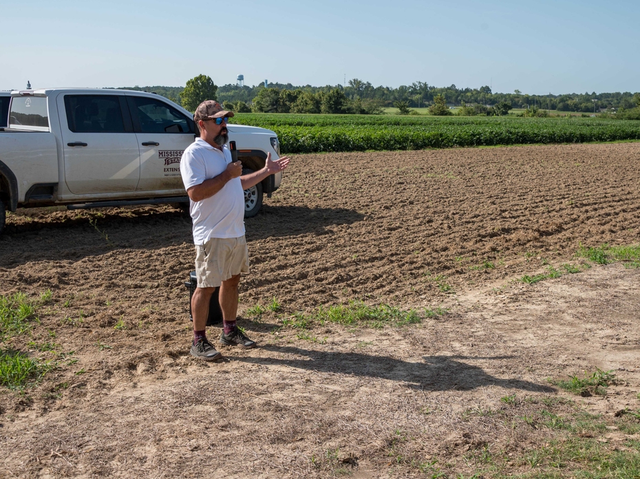 A speaker stands in front of research plots with a Mississippi State University Extension pickup truck in the background.