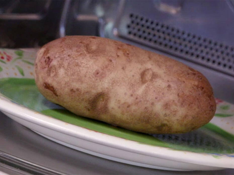 A close-up of a large brown Russet potato on a green plate with a white rim inside a microwave oven.
