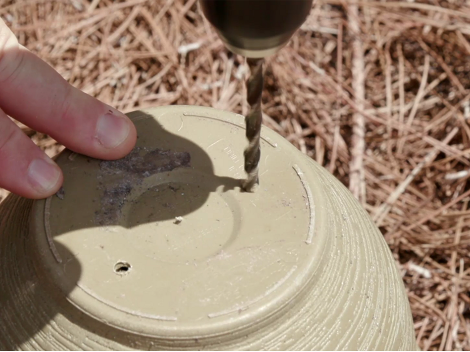 Fingers steady an upside-down flower pot as a drill bit pierces the bottom to make drainage holes. 