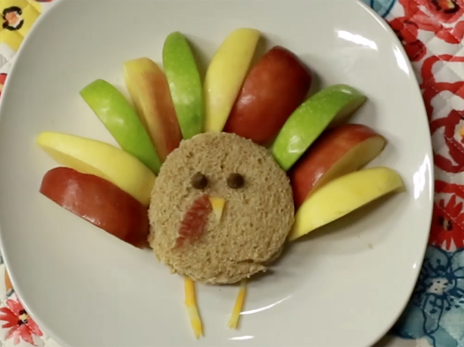 A round peanut butter and jelly sandwich sits on a white plate. It is decorated to look like a turkey, with green, red, and yellow apple slices for feathers, chocolate chips for eyes, cheese for a nose and legs, and a small bit of bacon for a wattle. The plate sits on a multicolored floral placemat. 