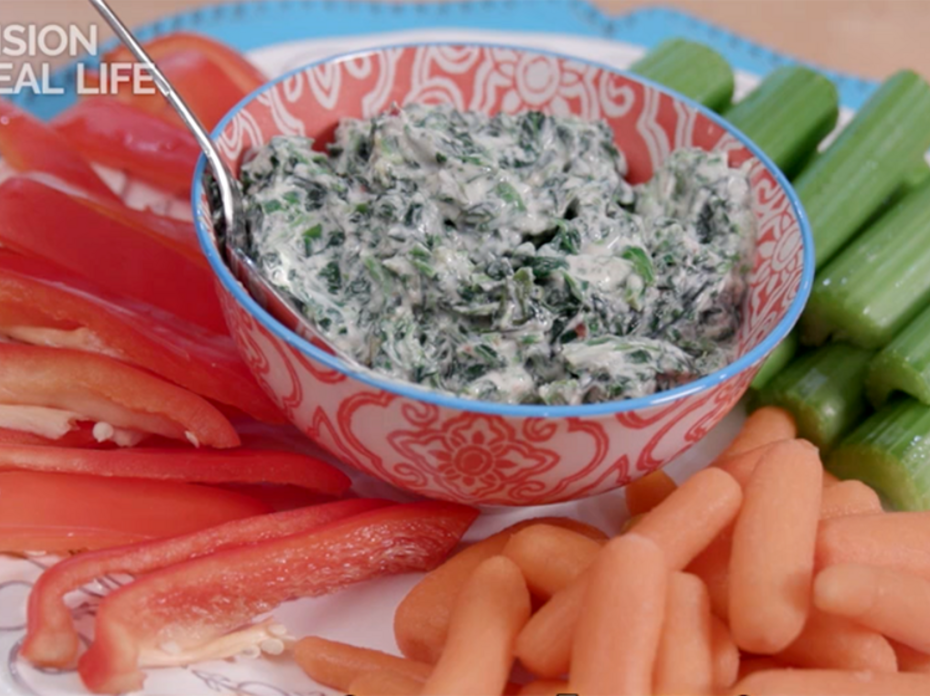 Spinach dip in a ceramic bowl centered on a plate surrounded by cut celery, baby carrots, and sliced red bell pepper.