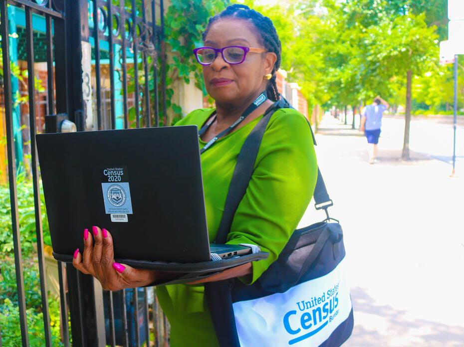 Woman in a green dress with a bag around her shoulder stands in front of a gate holding a laptop computer.
