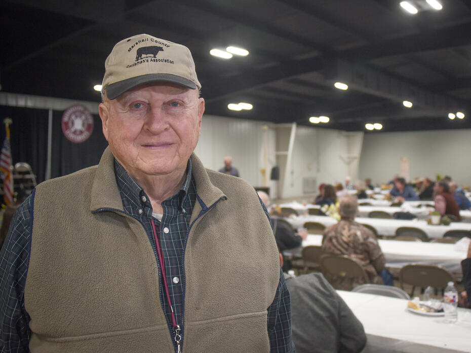 A man in a baseball cap stands in front of long tables with a crowd of people in the background.