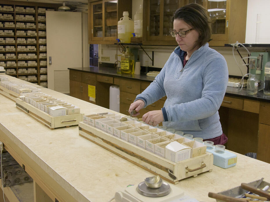 Small, cardboard containers in trays lie in a row on a workbench while a woman works with the contents of one.
