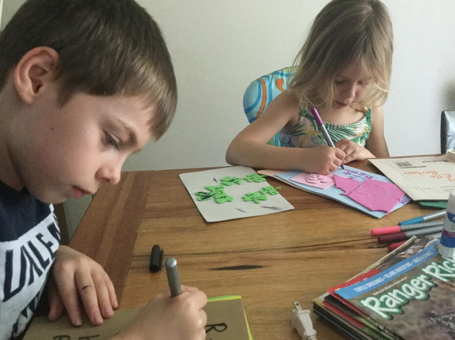 A boy and girl sit at a wooden table and use markers to draw on different colored sheets of paper.