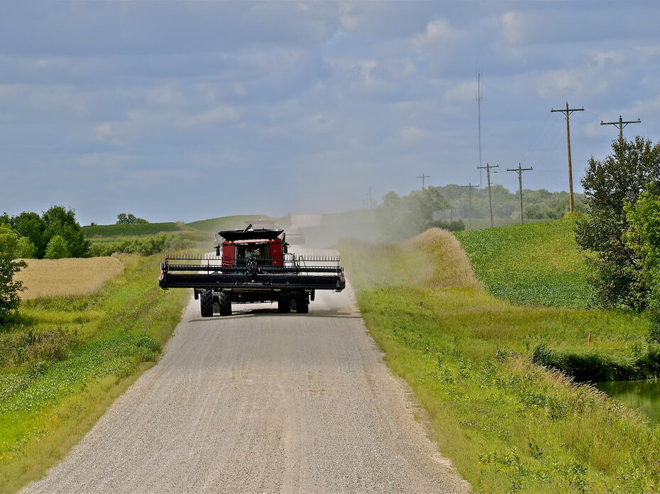 A red combine drives down a gravel road with farmland on both sides.