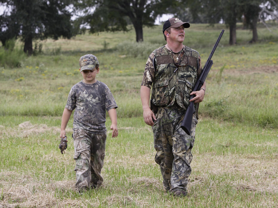 A man holding a shotgun and a boy dressed in camouflage walk in a grassy meadow.
