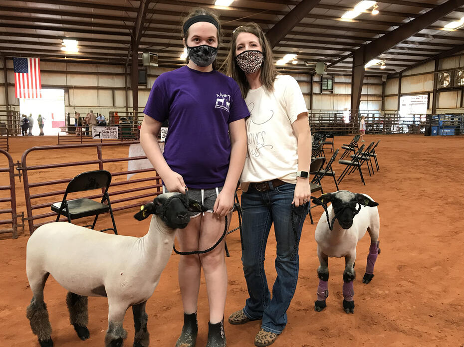 A daughter and her mother wearing masks in a rodeo building with two lambs leashed beside them.