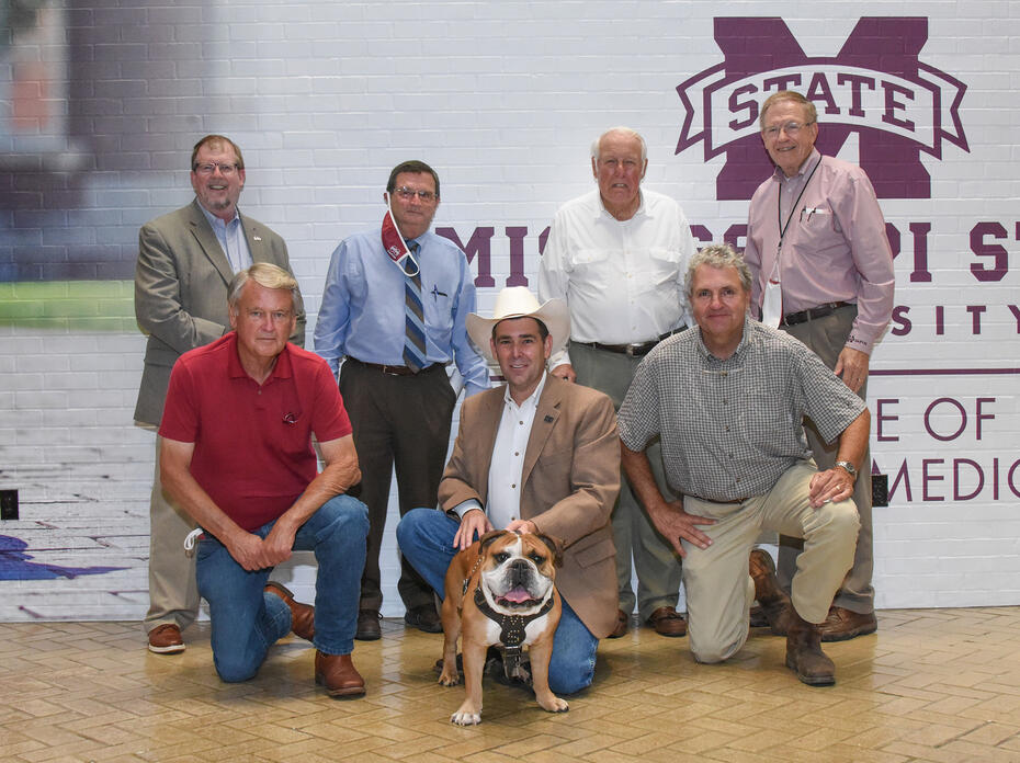 Seven men stand or kneel indoors in two rows, holding a bulldog in the center of the group.