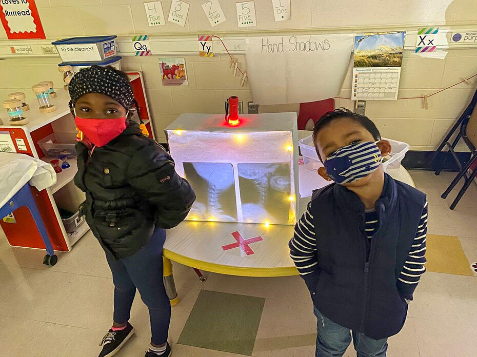 Two children in masks stand in front of an activity center in a classroom.