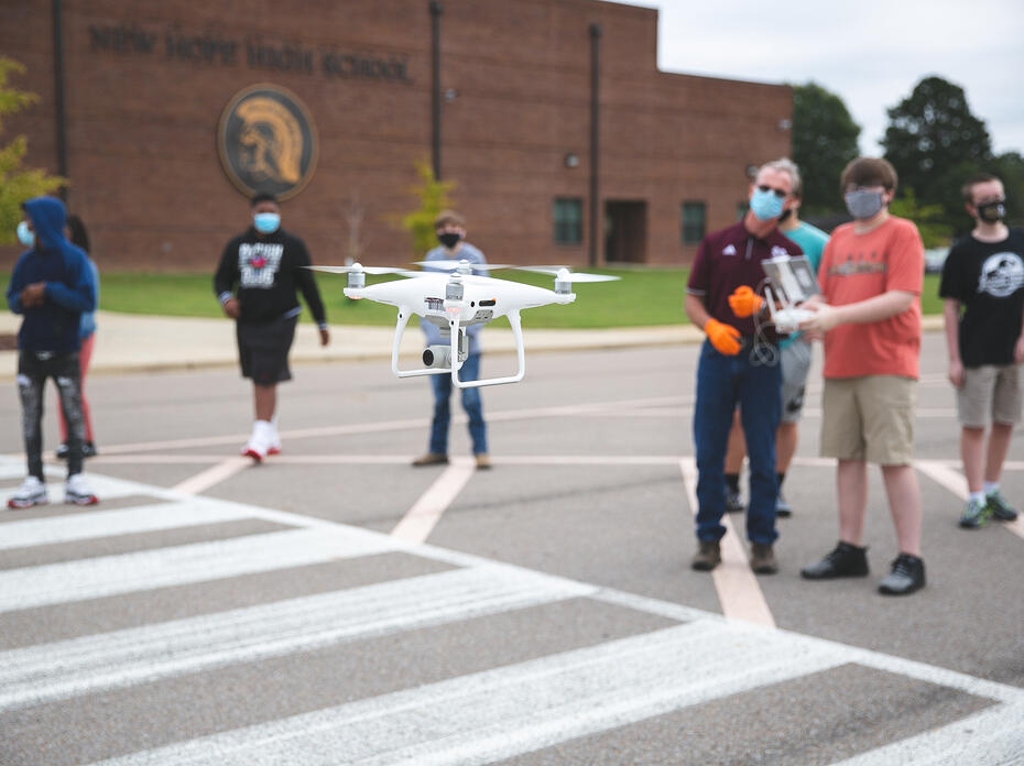 A drone in the foreground being controlled by young students in the background.