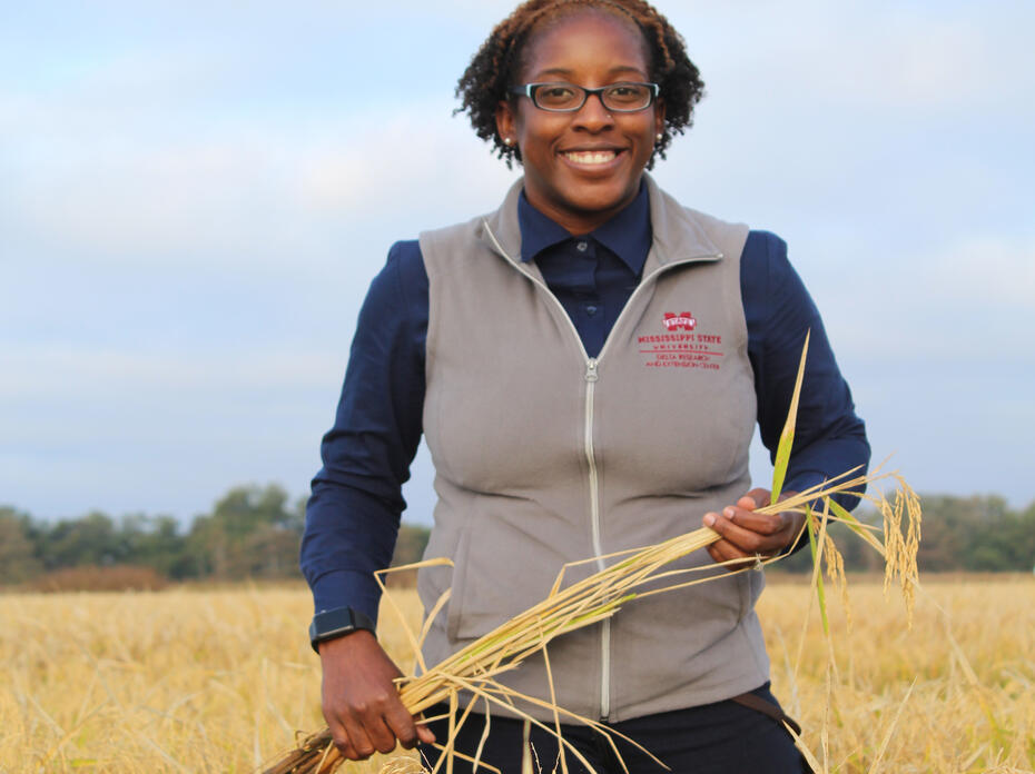 A woman holds a stalk of grain while standing in a field.