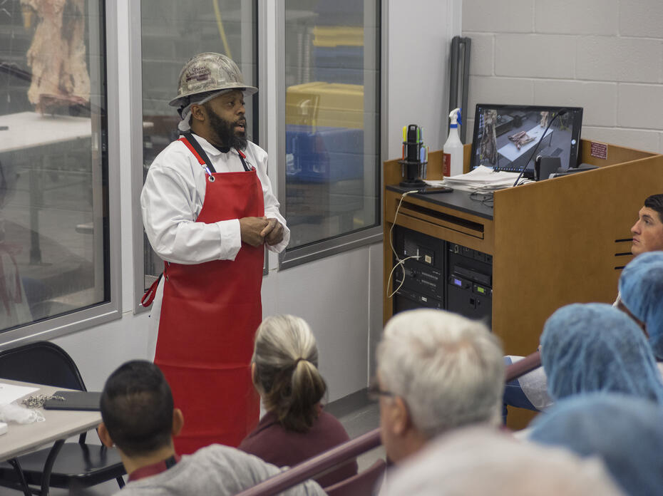 A man in a red apron speaks to a class in a lab.