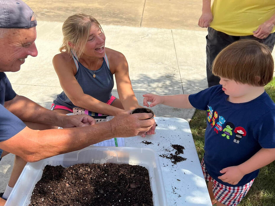 A man and a woman help a child plant seeds in a small pot of soil.
