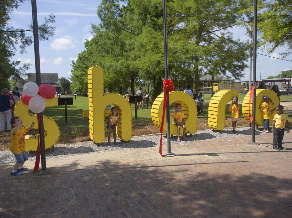 Children stand and sit around life-size, bright-yellow letters that spell out “share.”