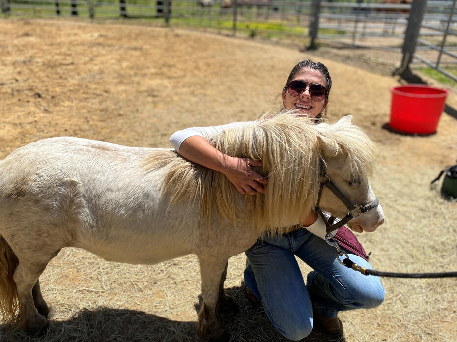 A woman kneels beside a small horse that she is hugging.