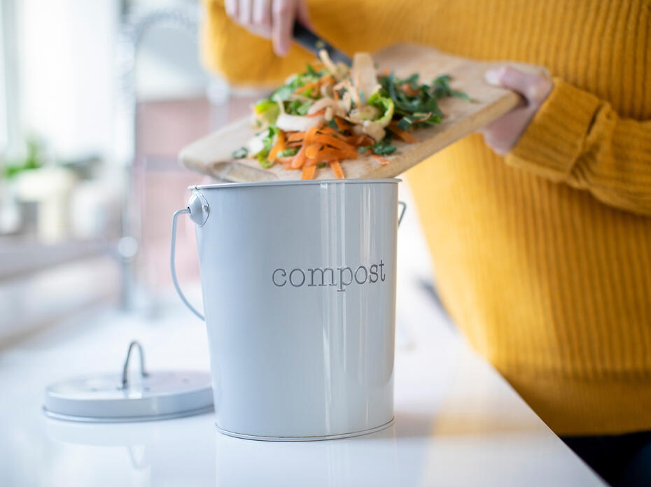 A woman scrapes vegetable scraps off a cutting board into a countertop compost bin.