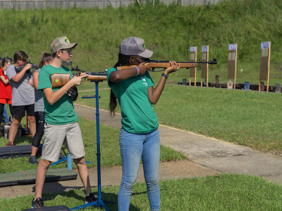 Teenagers at a shooting sports competition aim shotguns at targets.