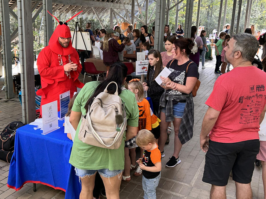 Several adults and children gather around an educational booth hosted by a man in a crawfish suit.