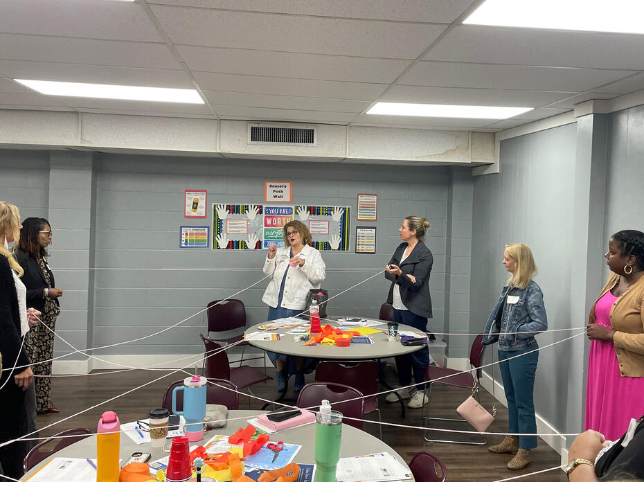 A group in a circle holds a white string that crisscrosses a classroom.