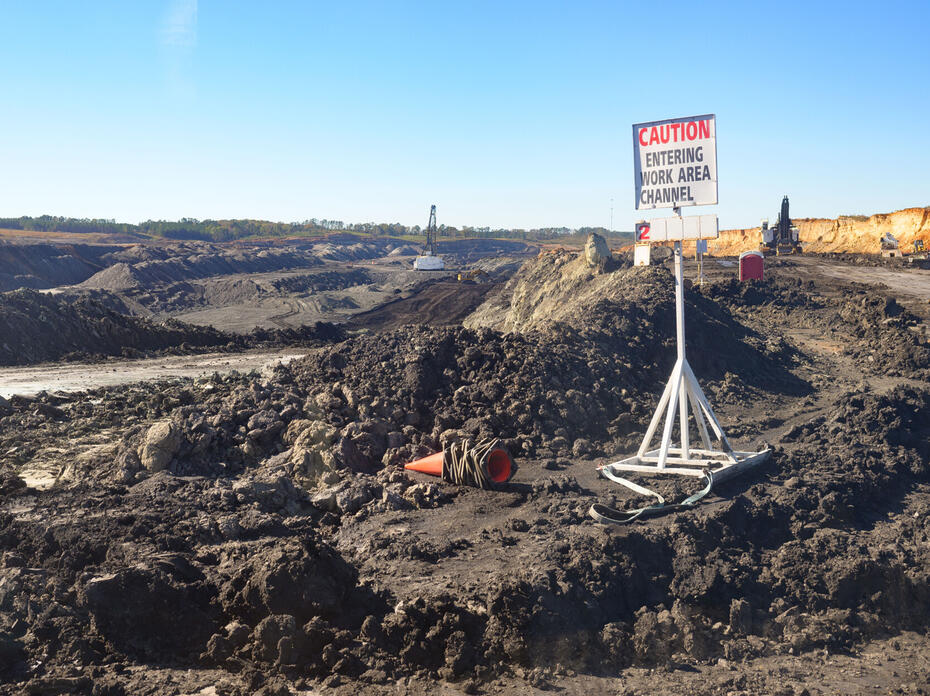 Wide shot of a surface coal mine featuring a layered landscape of exposed rock and soil, heavy machinery, and a sign that reads “caution, entering work area channel.