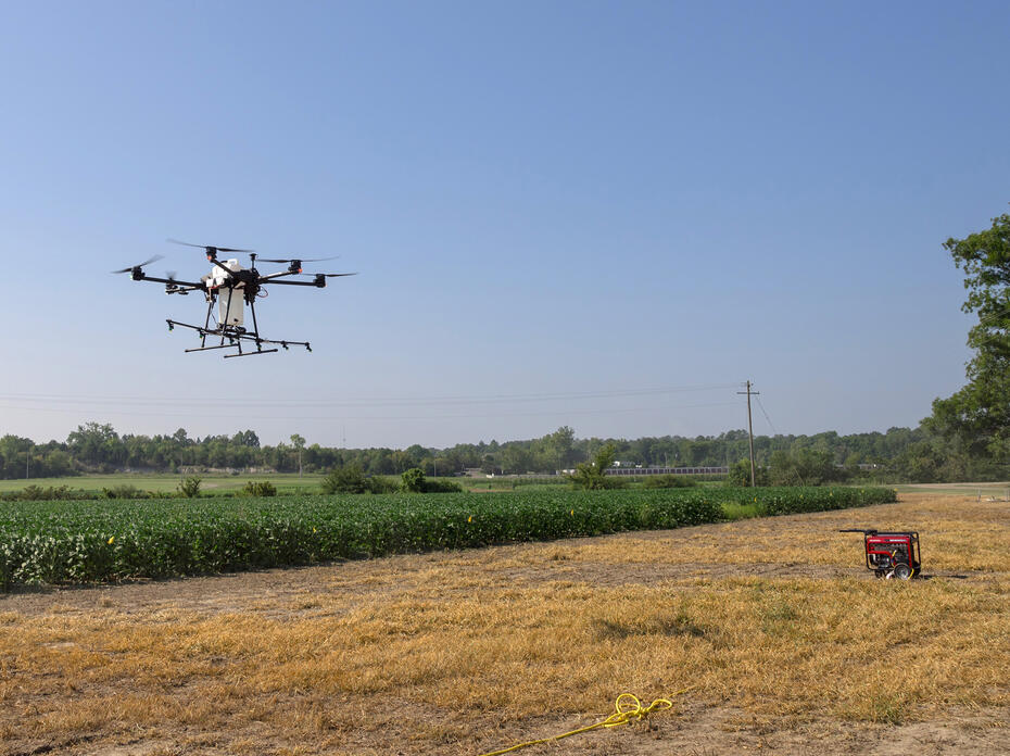 A large drone hovers over cropland.