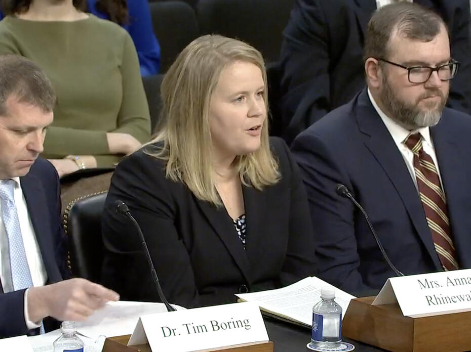 Woman seated and speaking into a microphone with men sitting beside her.