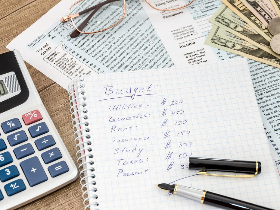 A calculator, handwritten budget, and paperwork sits on a table.