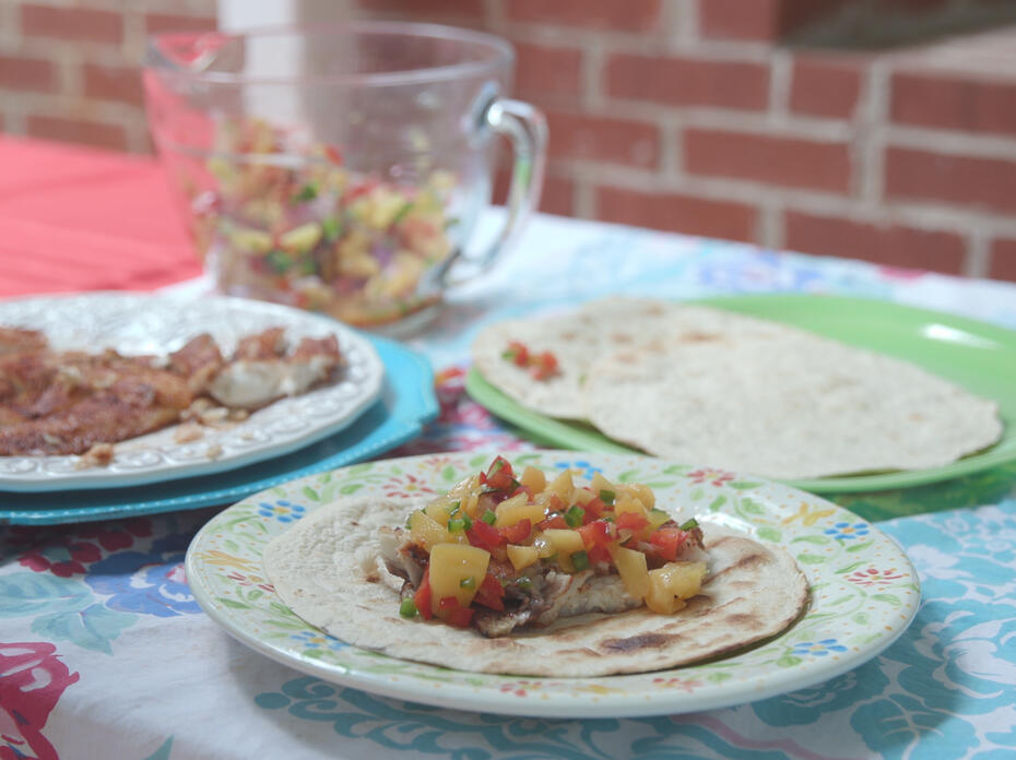 Three plates with tortillas, fish, and a completed grilled fish taco with peach salsa on a picnic table. 