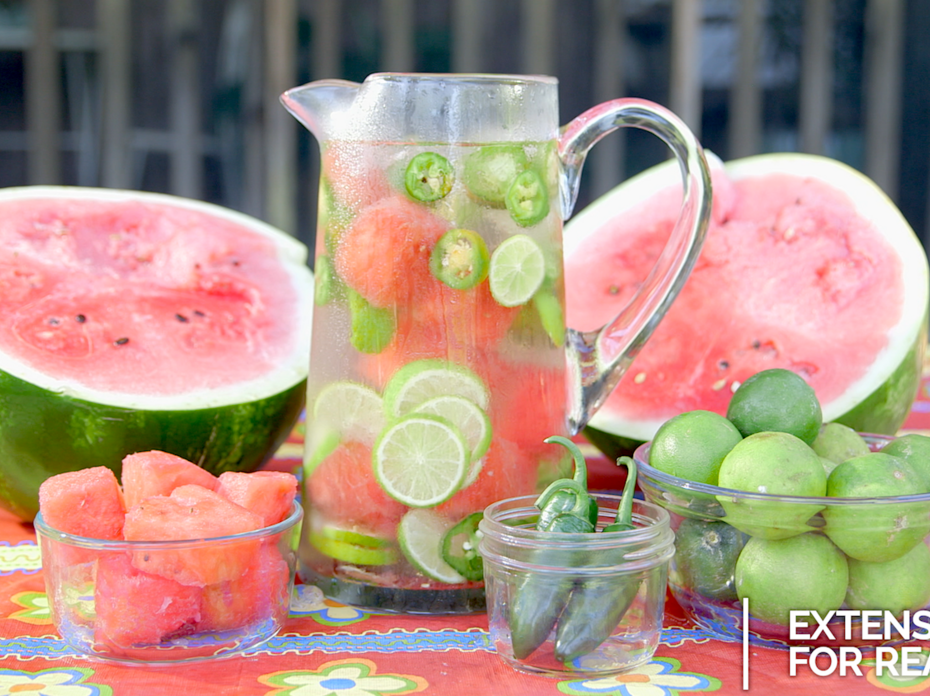 A pitcher of iced, flavored water sits in the center of a halved watermelon, bowls of cubed watermelon, limes and jalapenos.  