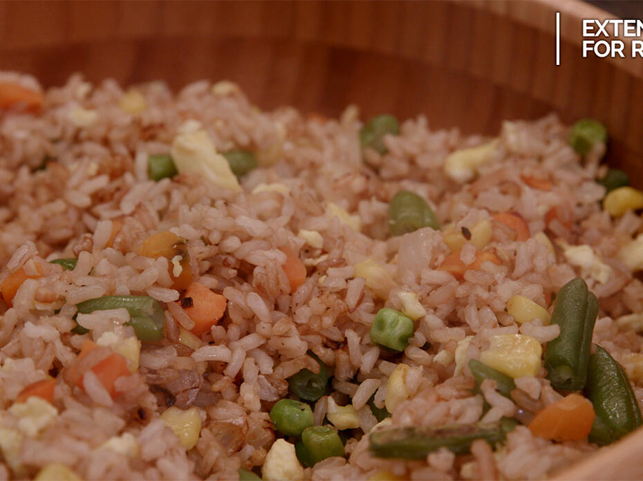 A brown bamboo bowl filled with fried rice made with brown rice, scrambled egg, and mixed vegetables including green beans, peas, carrots and corn. 